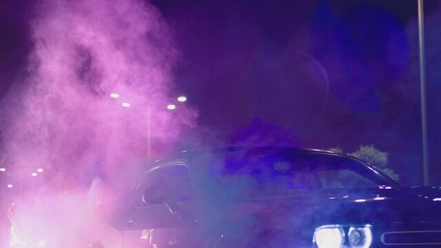 Young woman standing in middle of sport cars and waving , holding colorful smoke bombs flares . Girl starting muscle car drag racing at night . Back view slow motion . Ready for racing . Crowd of fans