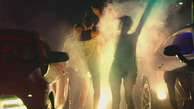 Young woman standing in middle of sport cars and waving , holding colorful smoke bombs flares . Girl starting muscle car drag racing at night . Back view slow motion . Ready for racing . Crowd of fans