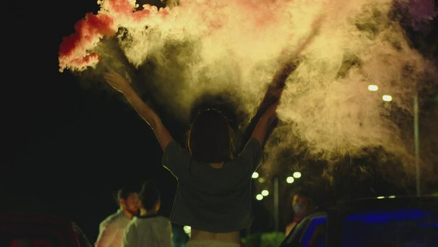 Young woman standing in middle of sport cars and waving , holding colorful smoke bombs flares . Girl starting muscle car drag racing at night . Back view slow motion . Ready for racing . Crowd of fans