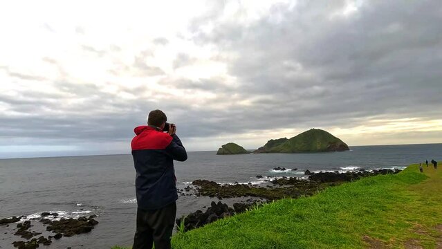Photographer taking pictures of the the cloudy horizon and the island called 'Ilhéu de Vila Franca do Campo' in the middle of the sea, in the island of Sao Miguel in Azores, surrounded of green grass