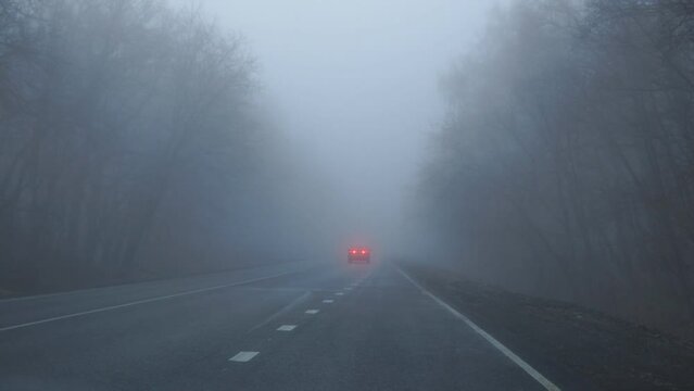 A car with its headlights on is driving in the fog along on an asphalt road