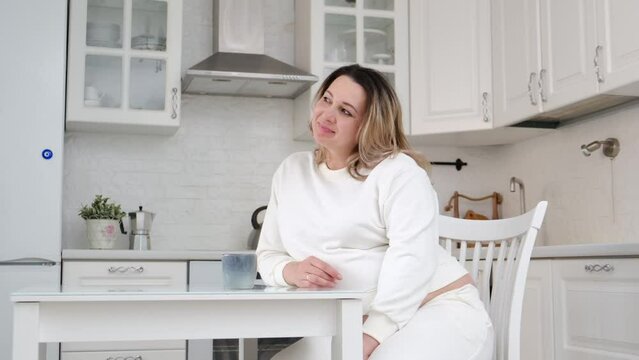 A pretty pergnant woman is drinking tea and sitting at table kitchen. Light background.