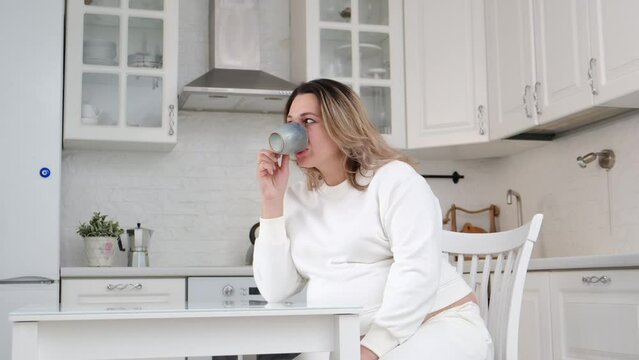 A pretty pergnant woman is drinking tea and sitting at table kitchen. Light background.