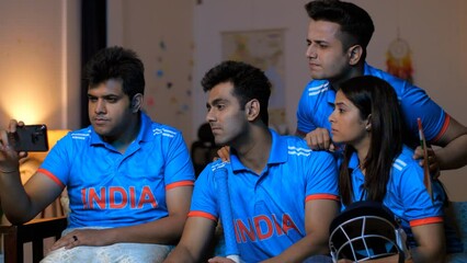 A pan shot of four Indian friends huddled together  watching a crucial cricket match on a smartphone  serious and tense expressions. Indian cricket team fans are engrossed in a match being broadcas...