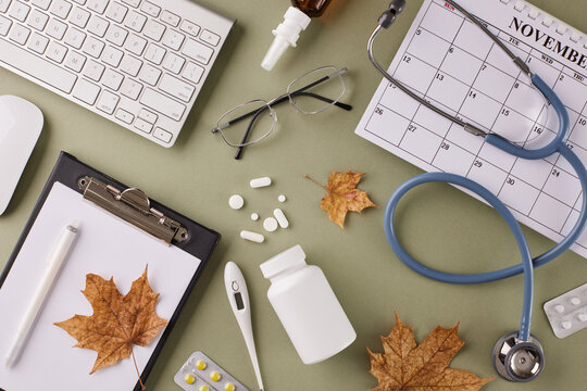 Connect from afar: remote medical consultation concept. Top view shot of keyboard, calendar, autumn leaves, eyewear, stethoscope, thermometer, tablets, clipboard, pen on green background