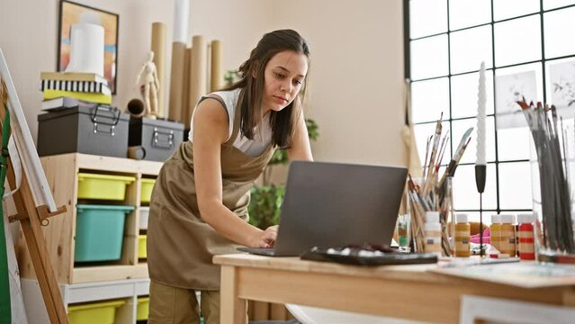 Bursting with creativity, young, beautiful hispanic woman artist standing proudly in her art studio, mastering her craft online using a laptop