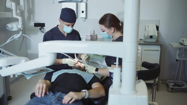 Male patient with opened mouth lying on dental chair while doctor and assistant doing dental treatment. Man having dental check-up in modern dentist office. Indoors