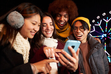 Group of multiracial friends smiling looking and pointing at cell phone outdoor at cold winter night. Four college students enjoying technologies using mobile standing together at amusement park. 