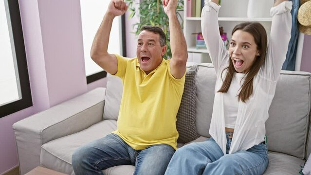 Hispanic father and daughter jubilantly celebrating a soccer match victory, watching tv together in their living room while resting on a comfortable sofa