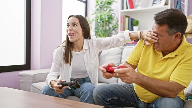 Hispanic father and daughter enjoy playing video game on the cozy home sofa, covering eyes and hands in laugh-filled skulduggery