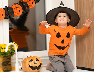 Halloween concept. A small, cheerful, handsome boy in a wizard's hat and an orange sweater sits on a table in the kitchen against the background of a pumpkin, a ghost and a bouquet of yellow flowers.