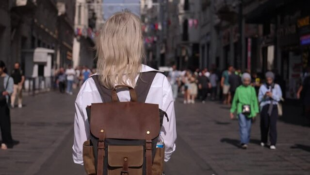 Back view of young beautiful blonde female tourist walking on street, summer fashion style, with backpack and white shirt, travel. Attractive girl exploring new city. Sunny Image Fashion Portrait