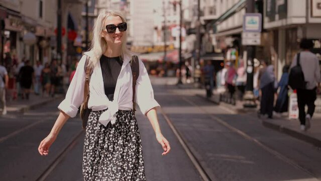 Young beautiful slender woman walking along busy city street. Tourist in stylish traveler's clothes, white shirt and backpack walks around sights and explores new places and streets.