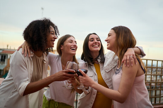 Group multiracial smiling female friends toasting glasses red wine and celebrating party outdoors. Young cheerful women hugging cheers on rooftop. Happy girls enjoying summer weekend vacation evening.