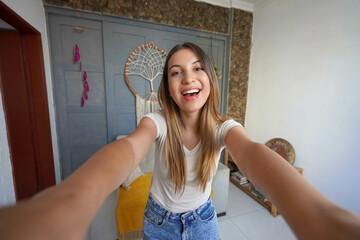 Happy young beautiful millennial girl taking a selfie smiling at the camera in the living room at...