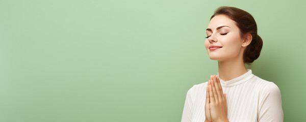 Young peaceful woman with closed eyes praying with folded hands in prayer gesture, blessing. Isolated on flat pastel green background with copy space, banner template. - obrazy, fototapety, plakaty