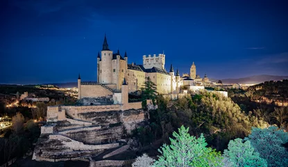Photo sur Plexiglas Cerro Torre Castillo medieval europeo sobre una colina por la noche, con algunas estrellas, vegetación en el primer plano y una ciudad también medieval al fondo, desde la ciudad de Segovia, España.