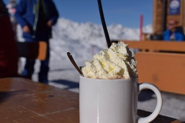Delicious hot chocolate outdoors in snowy ski resort. Fluffy whipped cream in blank white mug with snowcapped mountain background. Ideal hot drink for a rest during skiing and snowboarding holiday