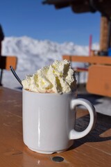 Delicious hot chocolate outdoors in snowy ski resort. Fluffy whipped cream in blank white mug with snowcapped mountain background. Ideal hot drink for a rest during skiing and snowboarding holiday