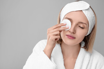 Young woman cleaning face with cotton pad on light grey background. Space for text