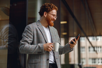 Handsome male freelancer holding phone while standing near office during break time and drink coffee
