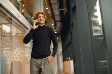 Smiling male sales manager is talking phone standing in modern coworking and looks away