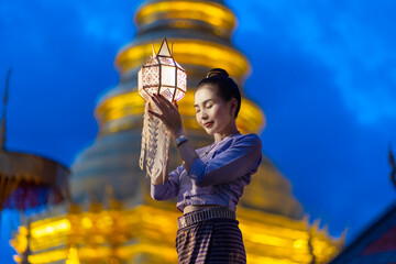 Asian woman wearing a traditional Lanna dress holding a colorful paper lantern raised high and praying to bless the holy things at night during Yi Peng lantern festival. Lamphun Province, Thailand © somchairakin