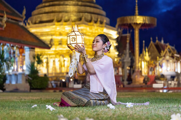 Asian woman wearing a traditional Lanna dress holding a colorful paper lantern raised high and praying to bless the holy things at night during Yi Peng lantern festival. Lamphun Province, Thailand