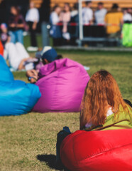 Fototapeta premium Crowded open air festival concert with musicians band on stage, rock show performance, with concert-goers attendees on beanbags, audience sitting on bean bag chair on a grass lawn on summer festival