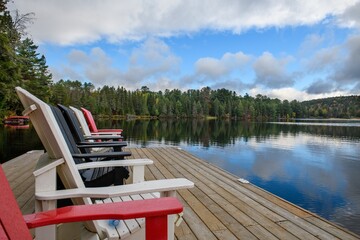 A few lounge chairs on a deck by the lake with autumn trees and reflections on the background in a beautiful sunny day