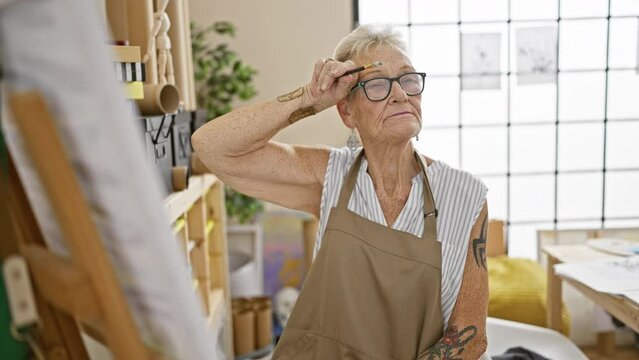 Sweating, yet smiling, the grey-haired senior woman artist confidently brandishing her paintbrush at the bustling art studio amidst a class of eager mature adults.