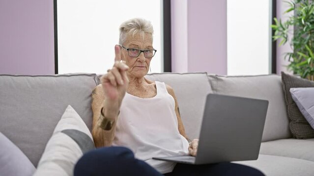 Serious grey-haired senior woman saying no with firm finger gesture while concentratedly using laptop on cozy home sofa