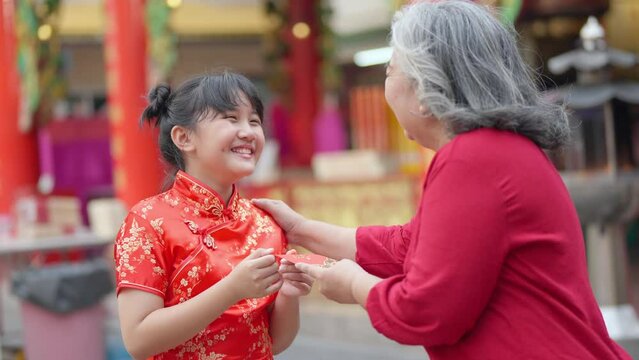 4K Happy Asian family grandmother giving red envelope contained money gift with blessing to little grandchild girl in Chinese red dress. Chinese lunar new year festival and holiday celebration concept