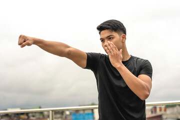 A young asian man makes a straight right hand while practicing his boxing skills on the rooftop of a building. Shadowboxing training, gritty urban scene. - obrazy, fototapety, plakaty