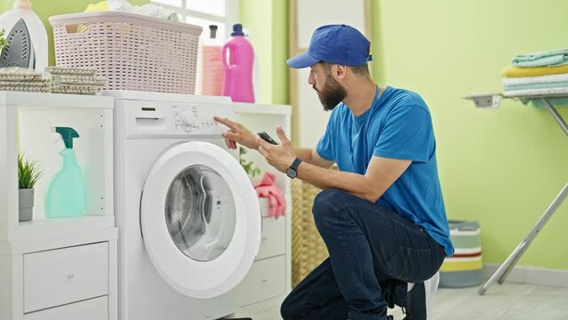 Young hispanic man technician repairing washing machine holding smartphone at laundry room