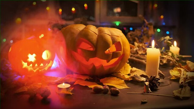 Two traditional carved pumpkins with sparkling candle inside lie on table among Halloween decorations of cones leaves and flickering candles and smoke reflecting colorful lights in dark.