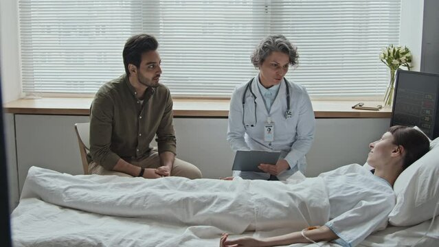 Female doctor talking with interracial couple about prospects of recovery and necessary treatment for woman lying in bed while her Middle Eastern boyfriend attentively listening to specialist