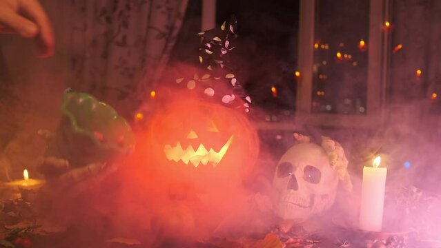 Male hand putting candy in a bucket Jack-o'-lantern and stroking skull among Halloween decorations in the fog and light of flickering candles, holiday background.