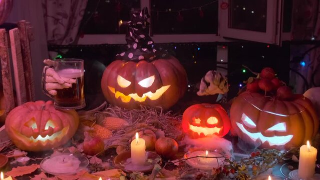 Glowing pumpkin with glass of beer slowly rotates head and waves his hand calling with his finger for Halloween party decorated with autumn holiday decorations in dry foliage with Jack-o'-lanterns.