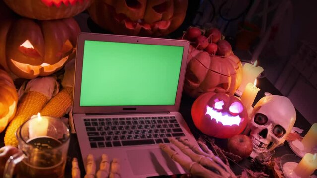 Creepy skeletal hands using a laptop with a green screen lying near Halloween decorations of blinking carved Jack-o'-lanterns in dark next to a skull and a glass of beer. Eerie scary spooky background