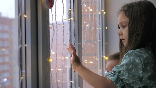 Adorable 5 years old girl sitting on the windowsill in home and looking through the window, admiring first snowflakes on street. Christmas time and miracle.Snow falling outside, celebrating New Year