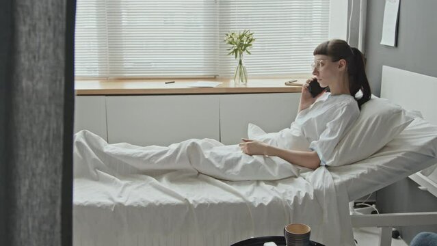 Panning shot of Caucasian female patient lying on bed in hospital ward talking over phone