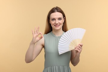 Happy woman with hand fan showing OK gesture on beige background