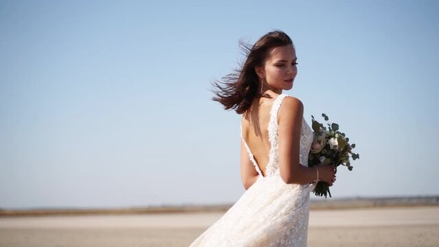 Happy young bride with bouquet of fresh flowers going at desert landscape. Elegance lady in gorgeous wedding dress walks in nature, windy weather. Pretty woman waits for a groom on wedding day.