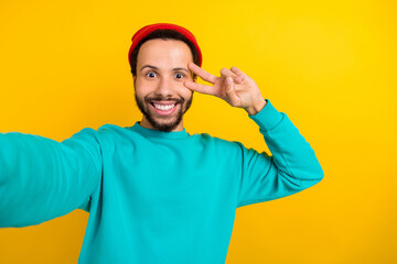 Portrait of positive funky man with beard dressed turquoise pullover making selfie show v-sign on eye isolated on yellow color background