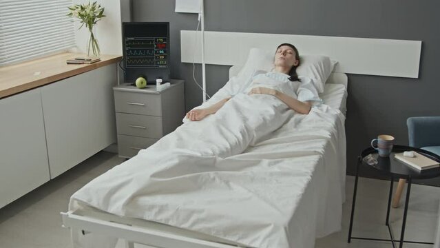 Long shot of Caucasian female patient on hospital bed connected with IV pole lying in coma in light clean medical ward