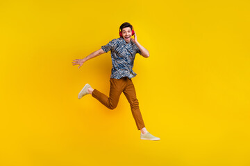 Fototapeta na wymiar Full size photo of eccentric guy wear stylish shirt pants flying touching headphones enjoy playlist isolated on yellow color background