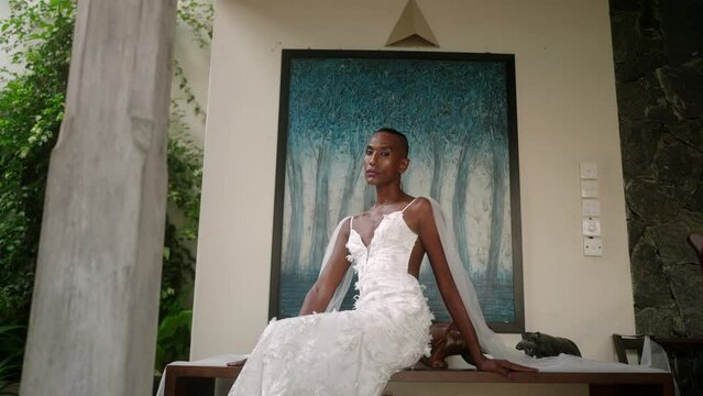 Gay black male in white wedding dress posing for fashion photo. Elegant gender fluid bride sits, turns to camera with abstract blue picture, trees on background in luxury retro villa. LGBTQIA