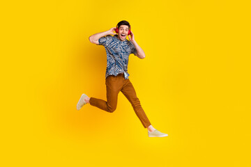 Fototapeta na wymiar Full size photo of impressed guy wear stylish shirt pants flying touching headphones listen music isolated on yellow color background