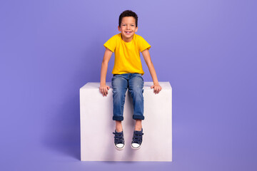 Full body photo of charming young boy sit white cube shopping promo dressed yellow outfit isolated on violet color background - obrazy, fototapety, plakaty
